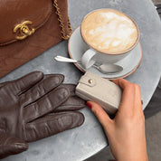 A person is holding a small, stone-colored travel jewellery box with a pair of brown leather gloves on top of it, in a café