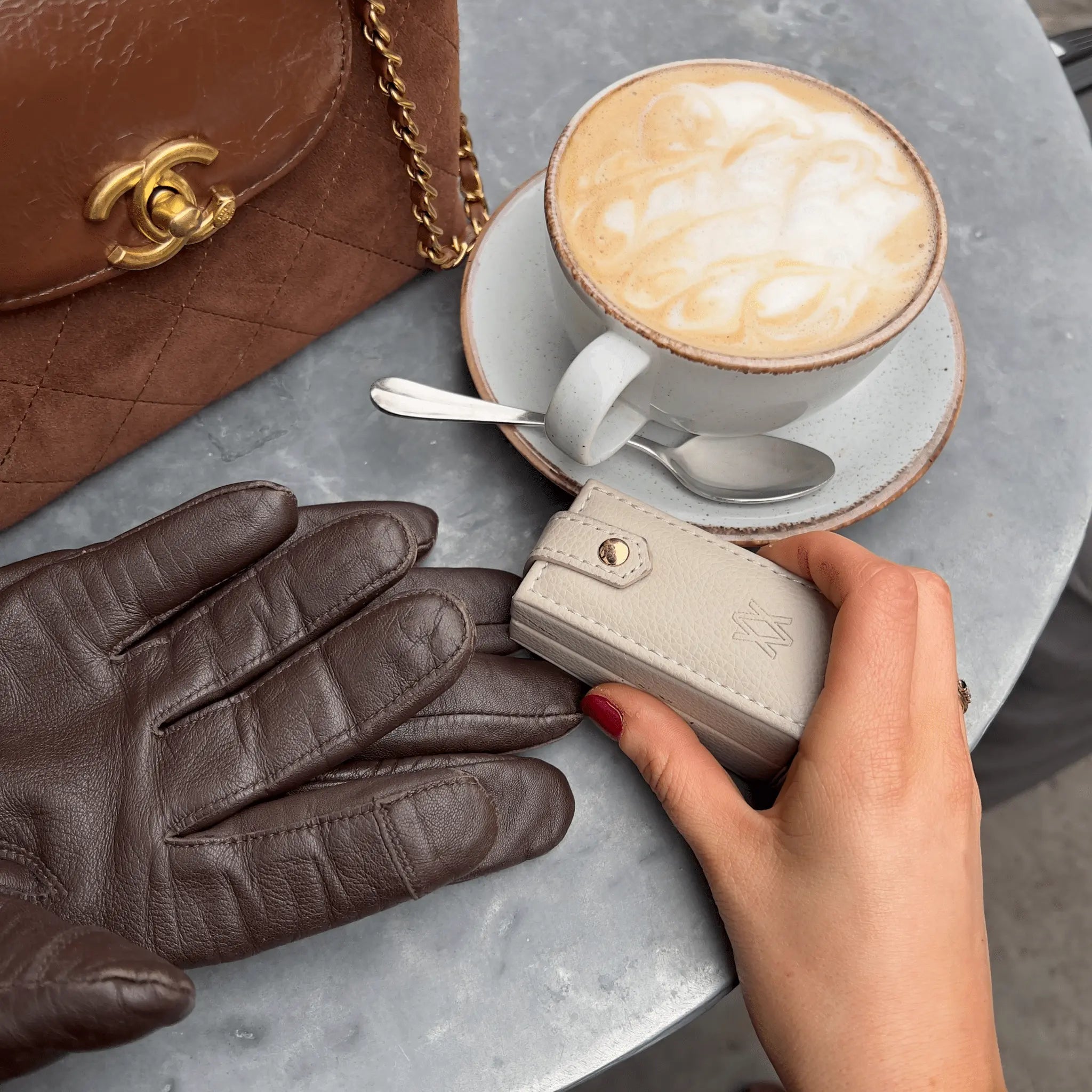 A person is holding a small, stone-colored travel jewellery box with a pair of brown leather gloves on top of it, in a café